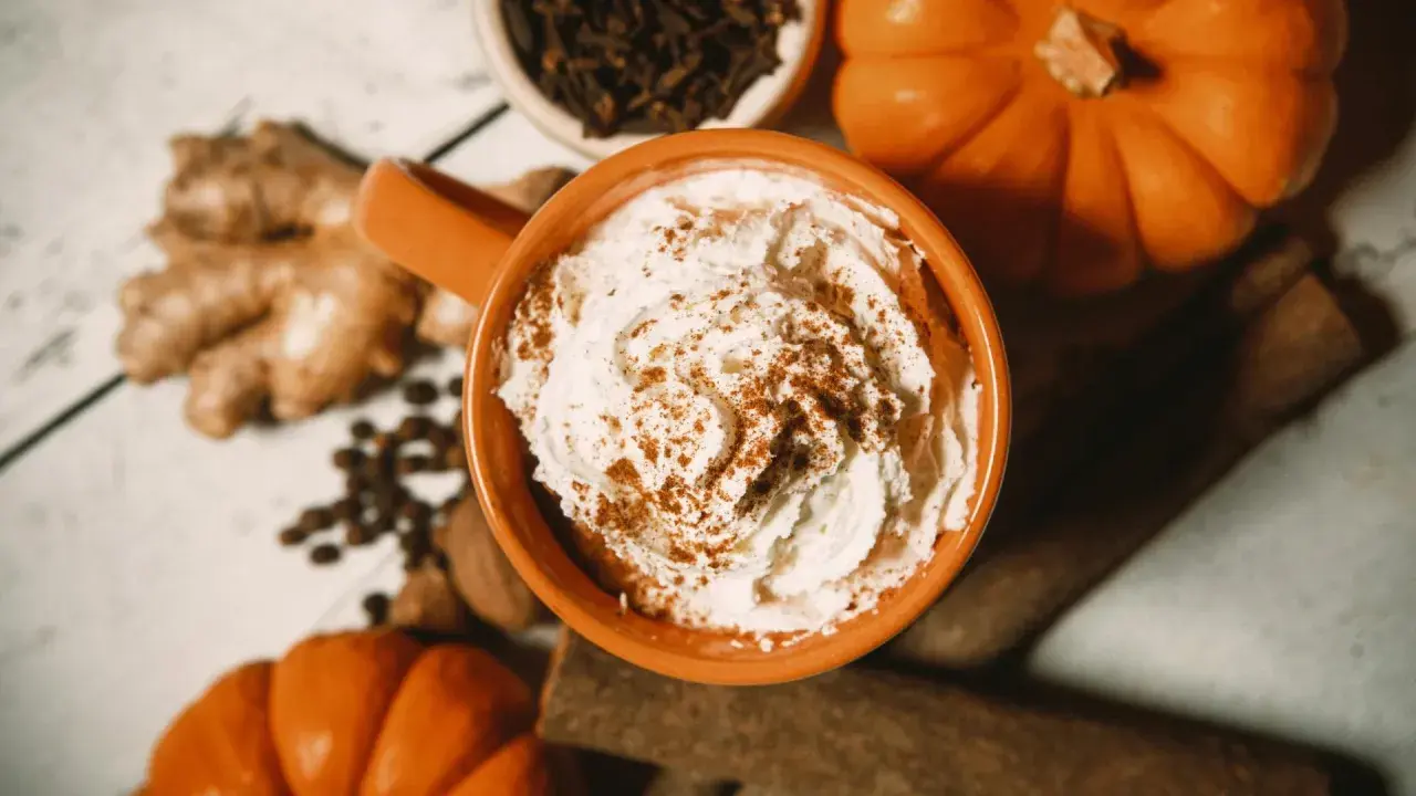Ein cremiger Pumpkin Spice Latte mit Sahne und Zimt, umgeben von Kürbissen, Ingwer und Gewürzen.