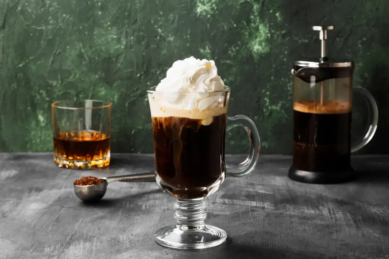 Ein Irish Coffee Rezept: Heißer Kaffee, Whiskey und Sahne in einem Glas, daneben eine French Press und ein Glas Whiskey.