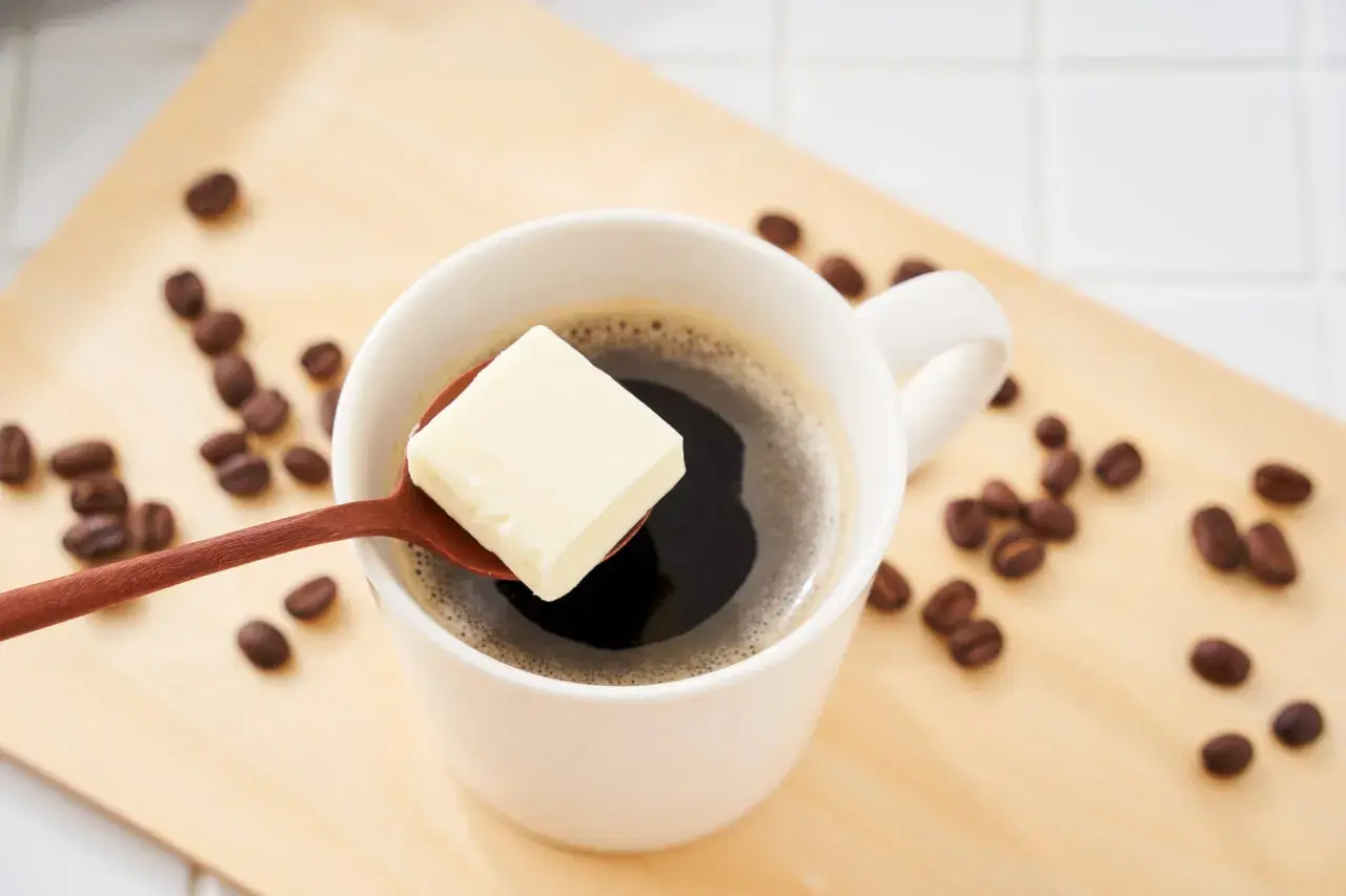 Ein Löffel Butter schwebt über einer Tasse Kaffee, umgeben von Kaffeebohnen auf einem Holztablett. Ein ungewöhnlicher Genuss: Kaffee mit Butter.