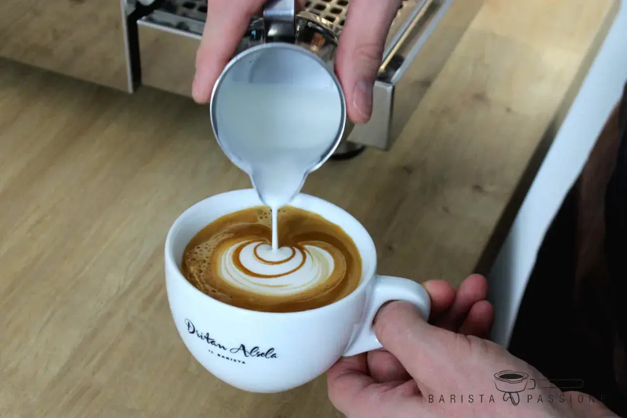 Barista vollendet die Flat White Zubereitung mit kunstvollem Latte Art Gießen. Milch fließt in Espresso und erzeugt ein Herzmuster.