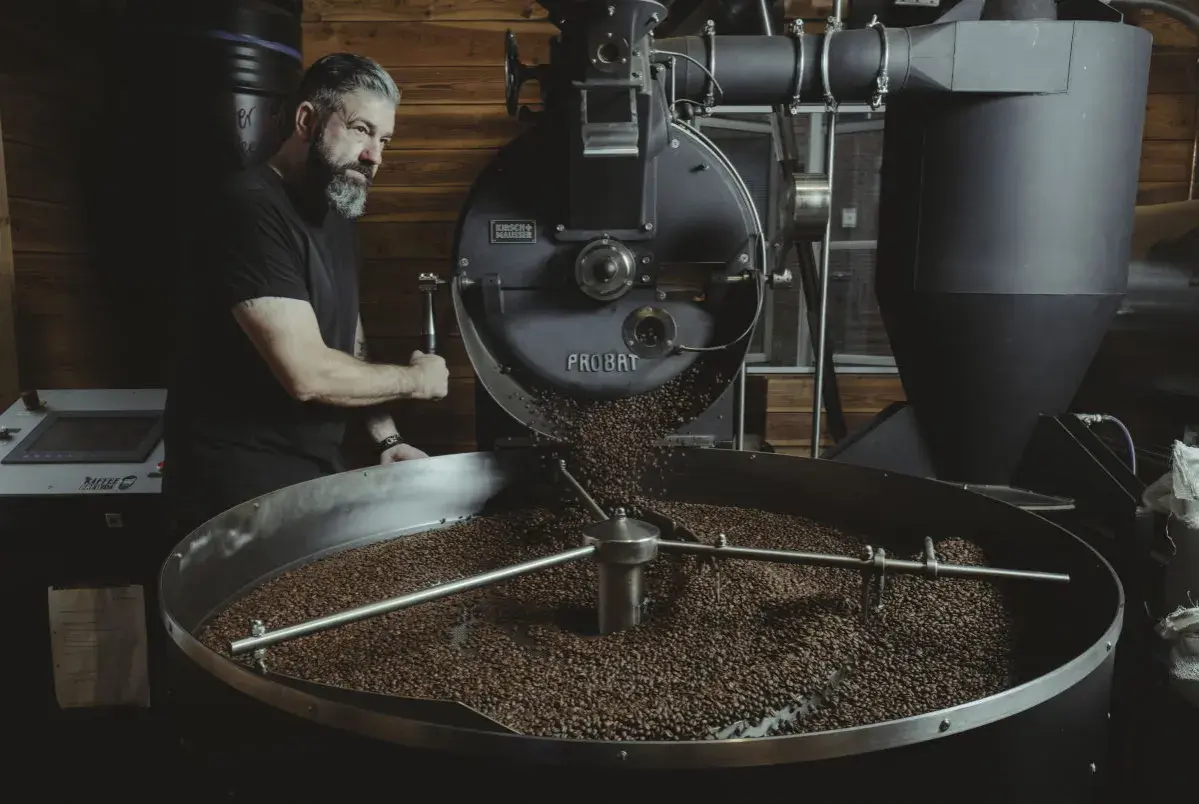 Mann mit Bart bedient Kaffeeröster. Frisch geröstete Bohnen rieseln in die Trommel. Ein Meisterwerk der dritten Welle.
