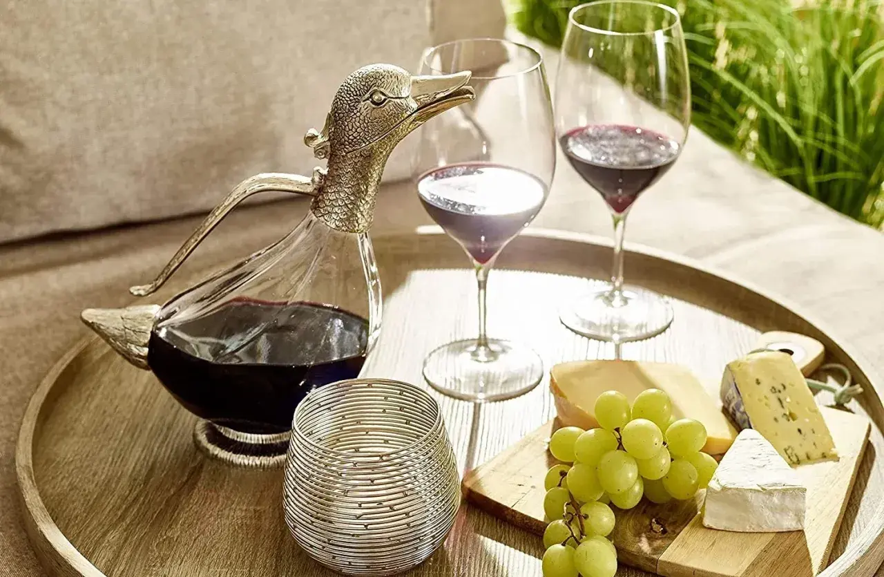 Ein Entenschnabel-Dekanter mit Rotwein, zwei Weingläser, Käse und Trauben auf einem Tablett – so gelingt das Wein richtig einschenken.