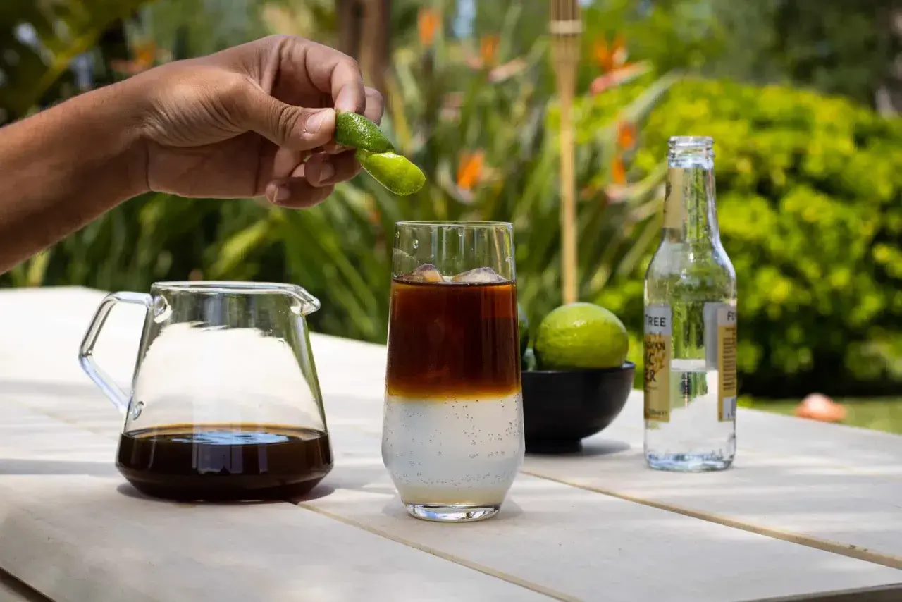 Hand presst Fingerlimette über ein Glas mit Eis, Kaffee und Tonic Water. Ein Krug mit Cold Brew und eine Flasche Tonic stehen daneben.