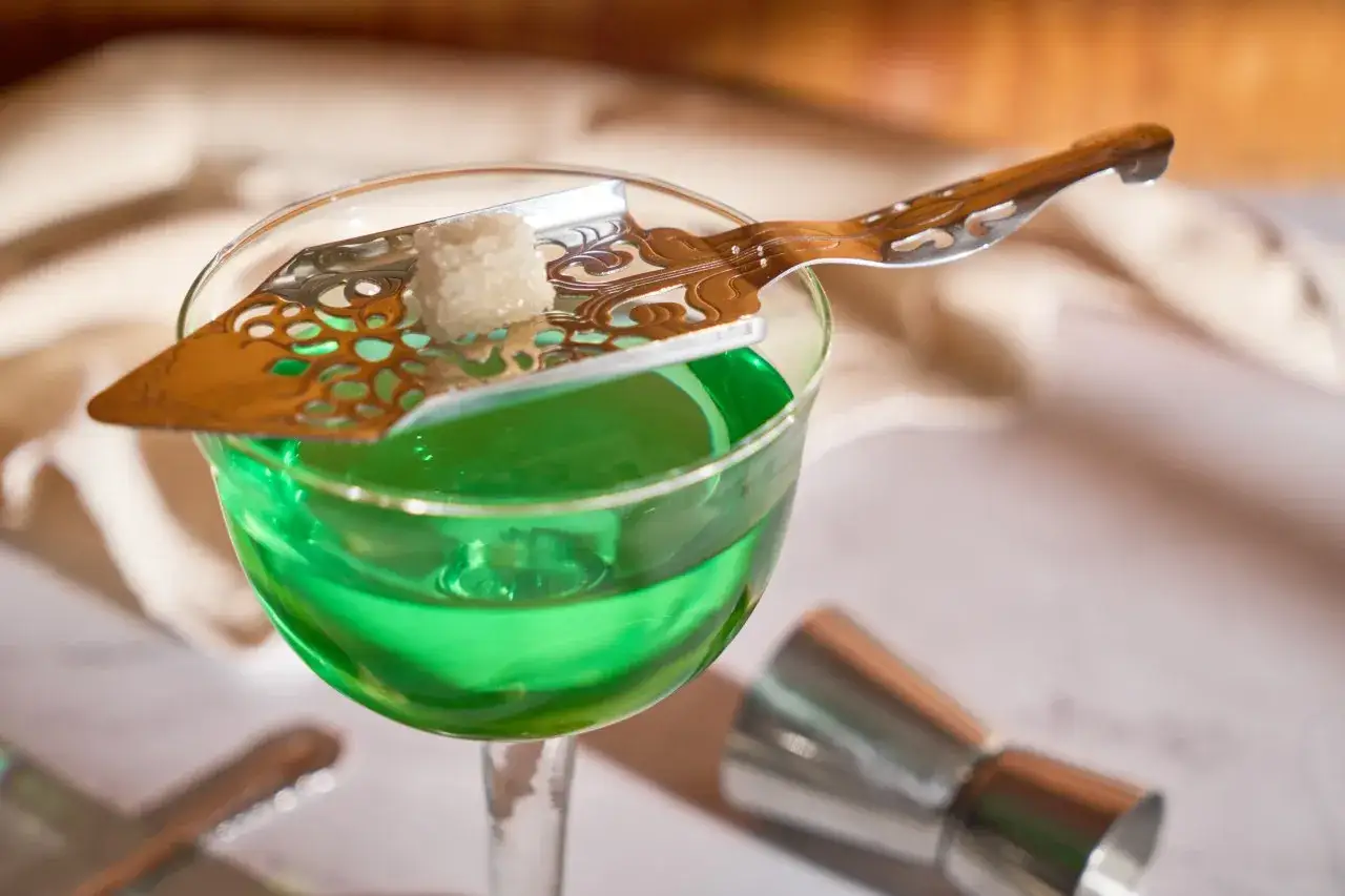Ein Glas mit leuchtend gr&uuml;nem Absinth, einem Branntwein aus Wermut, bereit zur Zubereitung mit einem Zuckersirup-L&ouml;ffel.