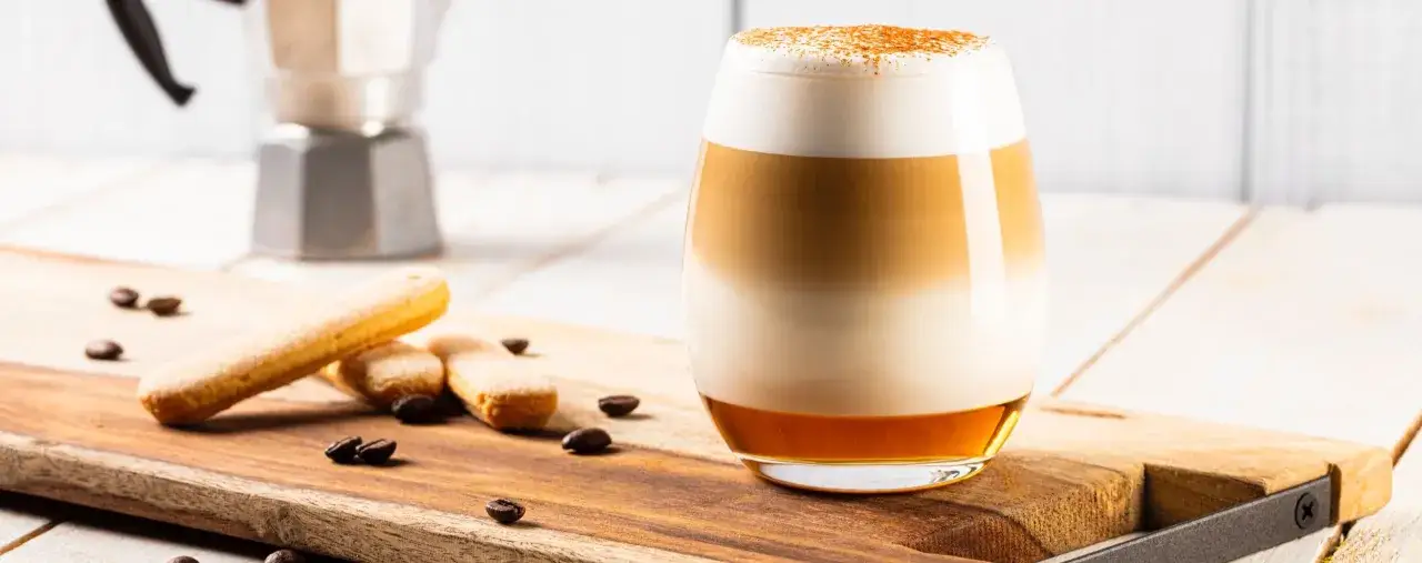 Ein geschichteter Spice Latte mit Milchschaum und Zimt, daneben Löffelbiskuits und Kaffeebohnen auf einem Holzbrett.