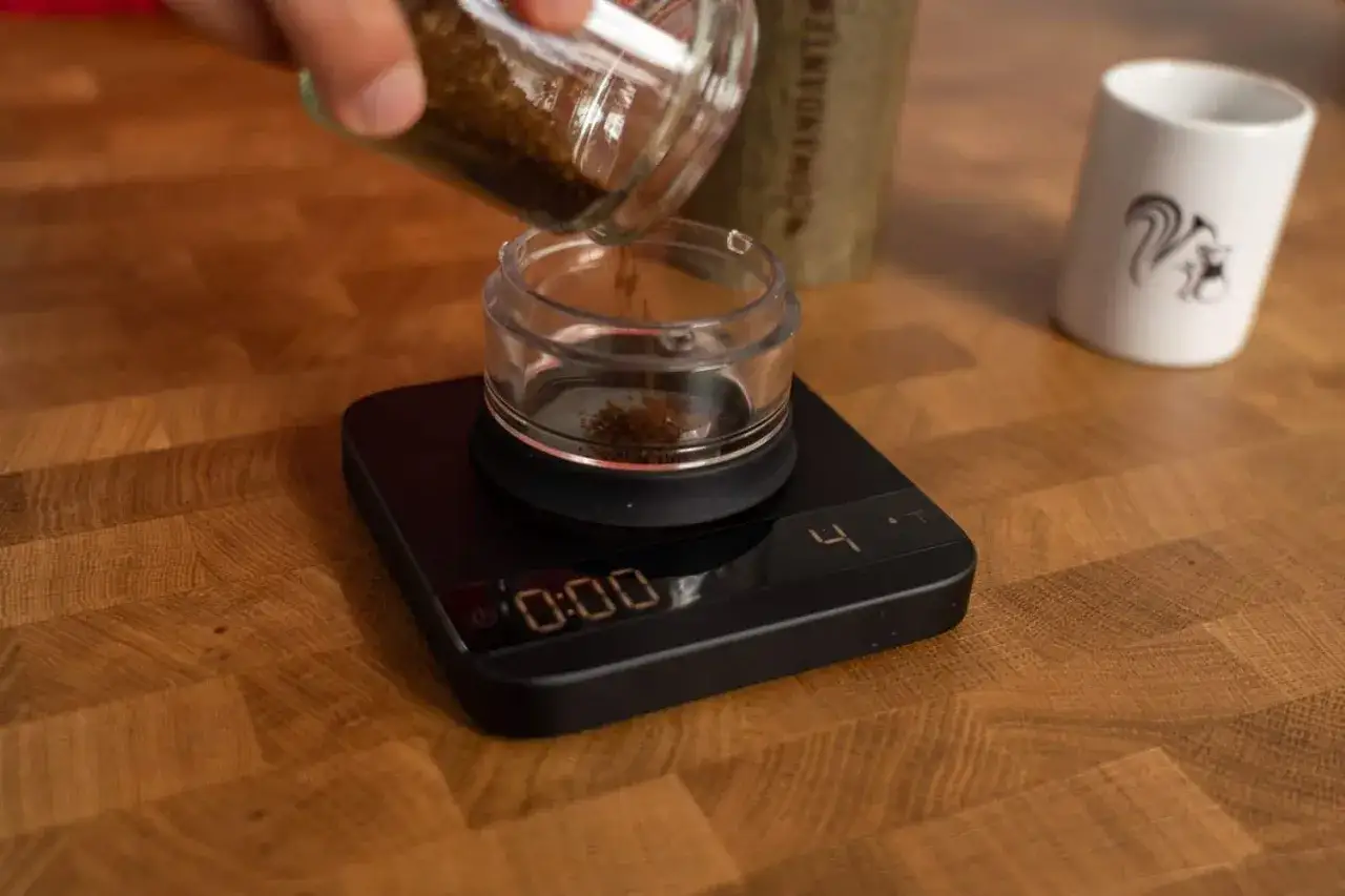 Kaffee für French Press wird abgewogen. Eine kleine Menge gemahlener Kaffee rieselt aus einem Glasbehälter auf eine digitale Waage.