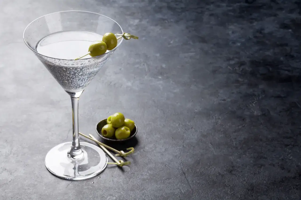 Ein Glas mit einem klaren Drink, garniert mit zwei Oliven, steht auf einem dunklen Tisch. Daneben eine kleine Schale mit weiteren Oliven. Ein klassischer trockener Wermut.