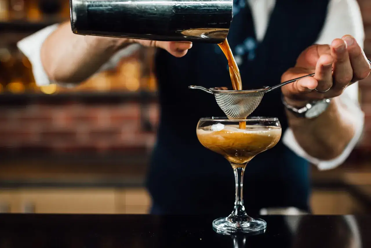 Ein Barkeeper mixt einen Espresso-Cocktail. Der dunkle, schaumige Drink wird durch ein Sieb in ein Glas gegossen.