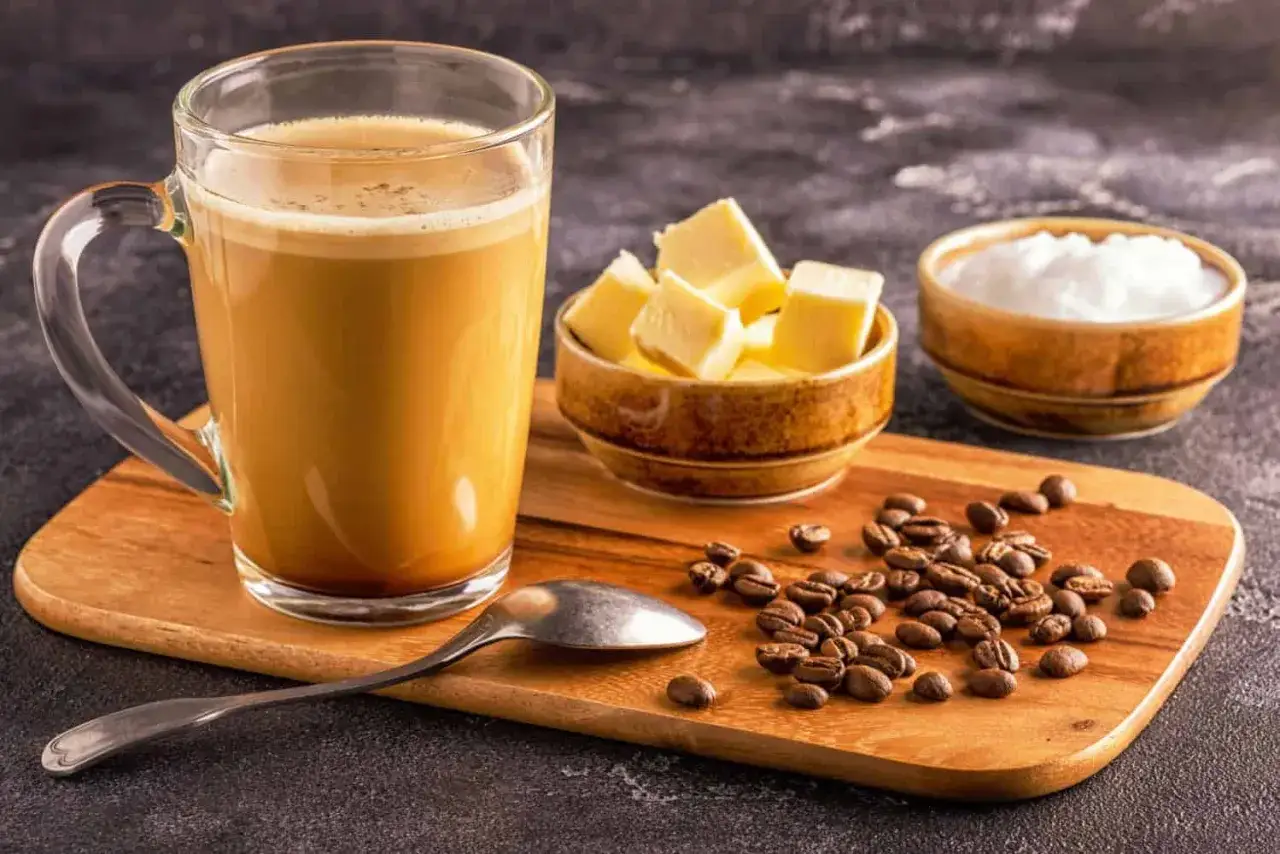 Ein Glas cremiger Kaffee mit Butter, daneben eine Schale mit Butterw&uuml;rfeln, Kaffeebohnen und Zucker.