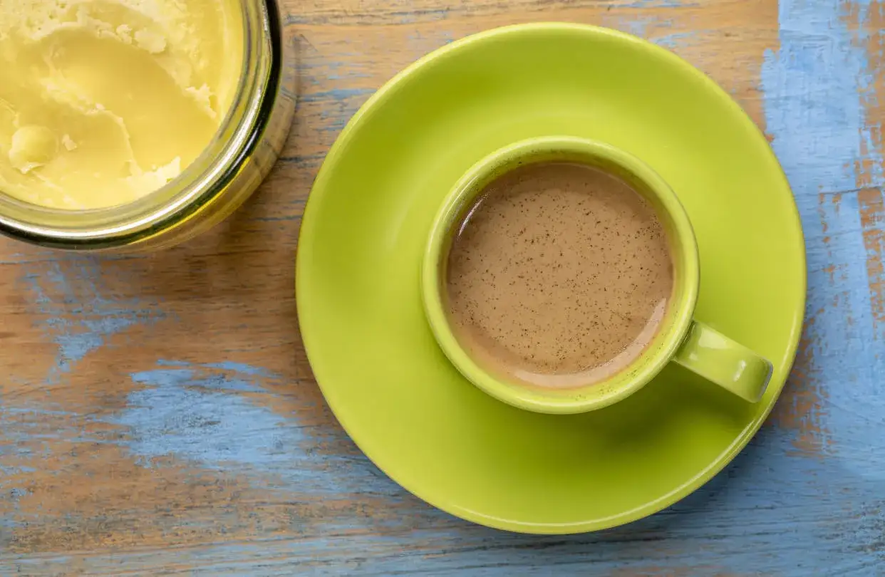 Kaffee mit Butter zum Abnehmen: Eine Tasse cremiger Kaffee neben einem Glas Ghee auf einem rustikalen Holztisch.