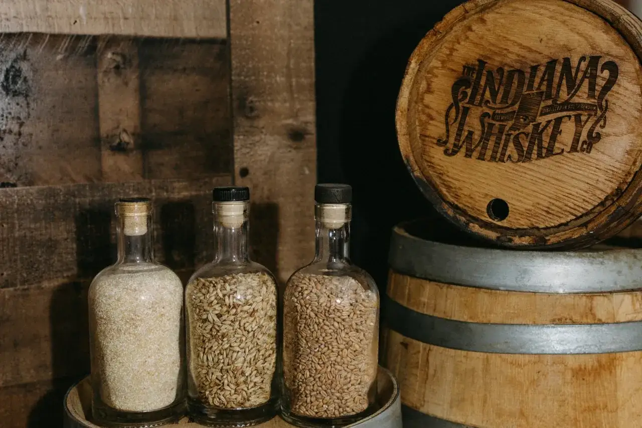 Drei Flaschen mit Malz und Gerste stehen vor einem Whiskyfass. Die Frage, ob Whisky glutenfrei ist, wird hier durch die Zutaten beantwortet.
