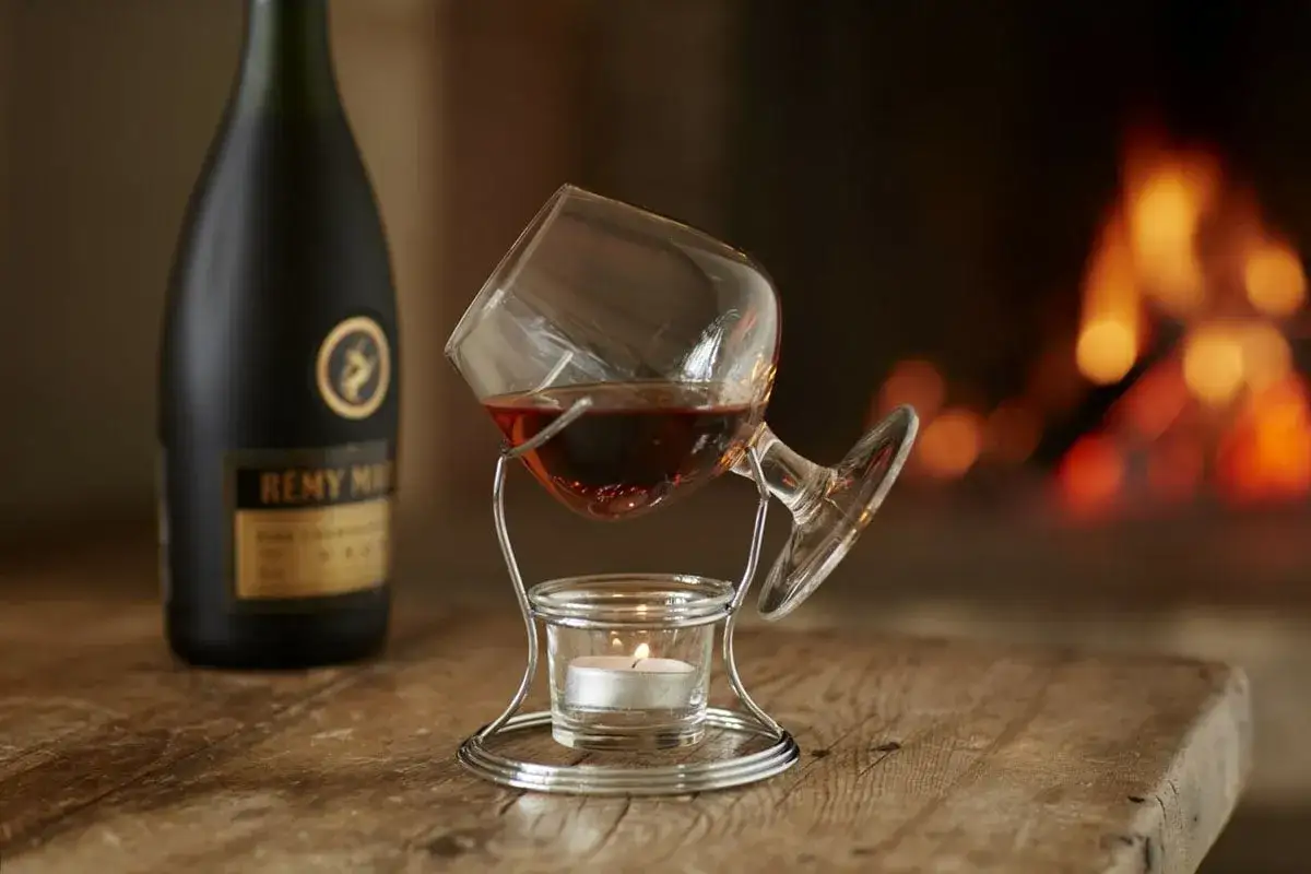 Ein Glas mit bernsteinfarbenem Brandy Cognac wird über einem Teelicht erwärmt, daneben eine Flasche Remy Martin. Gemütlicher Abend am Kamin.