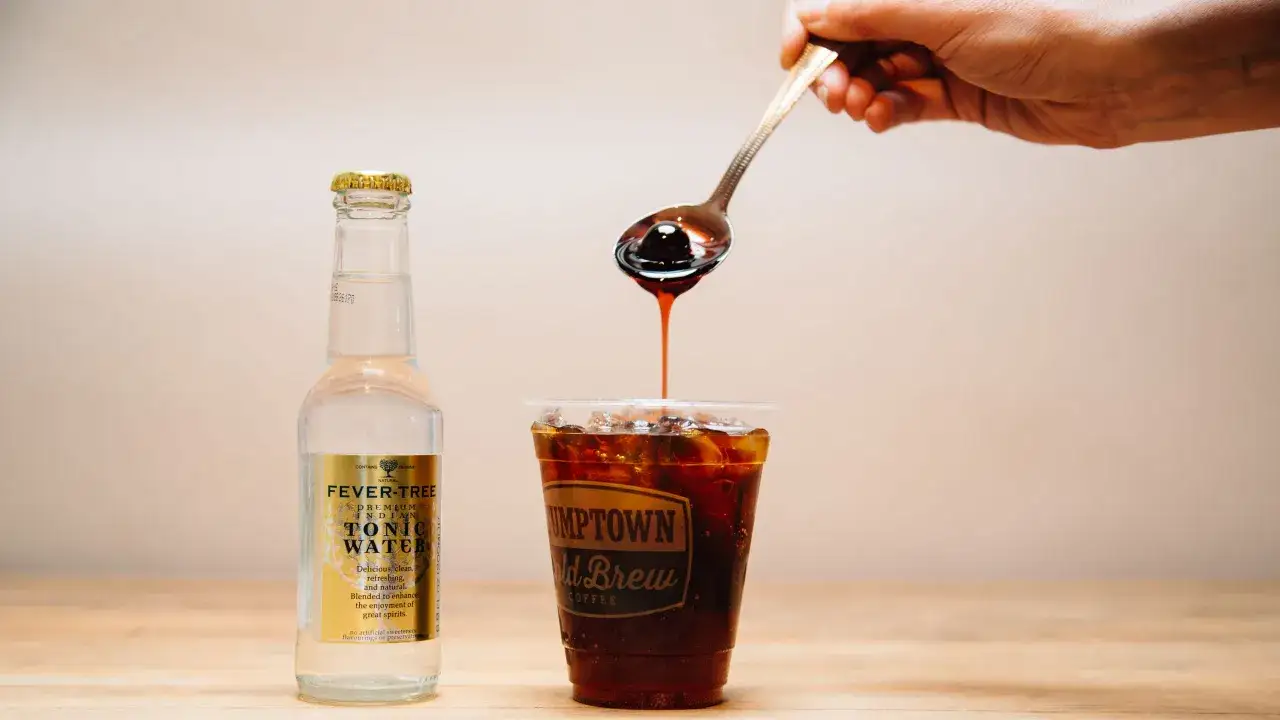 Ein Löffel dunkler Sirup tropft in ein Glas mit Eis und Kaffee. Daneben steht eine Flasche Tonic Water. Perfekt für einen erfrischenden Cold Brew Tonic.