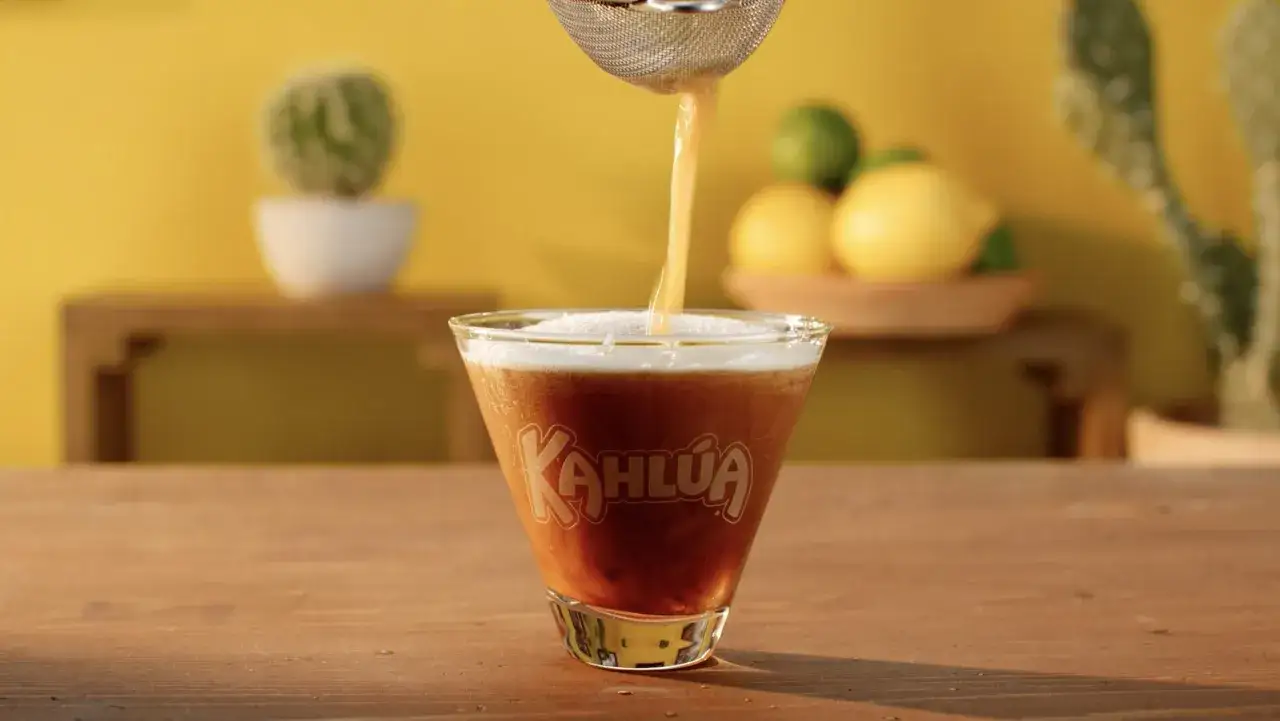 Ein Kahlúa Cold Brew Cocktail wird in ein Glas gesiebt. Im Hintergrund sind Kakteen und Zitrusfrüchte zu sehen.