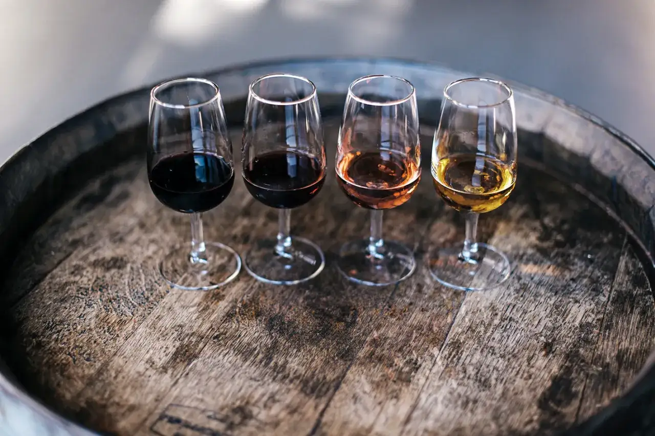 Vier Gläser mit unterschiedlichen Weinen, von dunkelrot bis goldgelb, stehen auf einem alten Holzfass. Ein Vergleich von Portwein und Sherry.