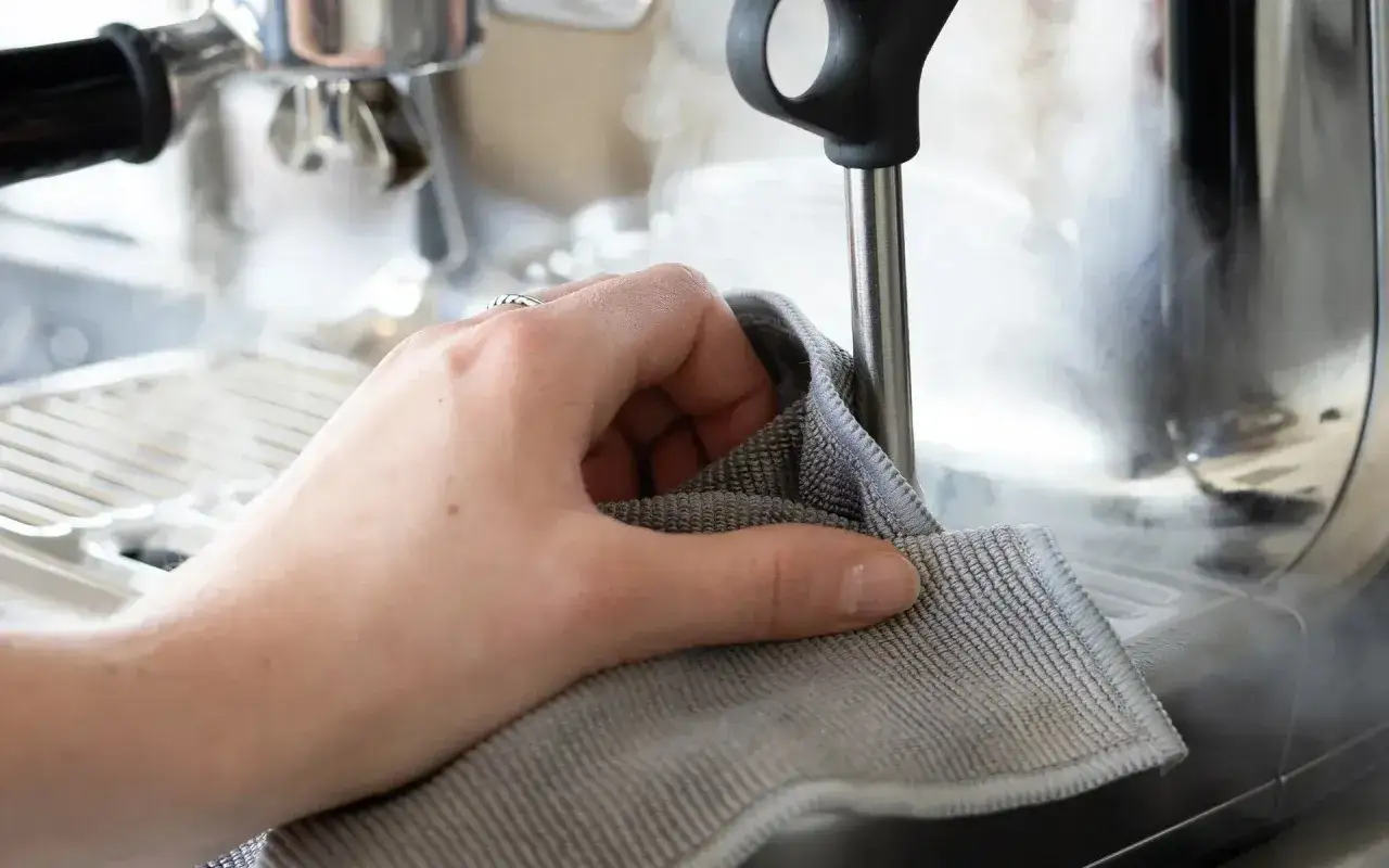 Hand wischt mit grauem Tuch über dampfende Espressomaschine. Innenreinigung für perfekten Kaffeegenuss.