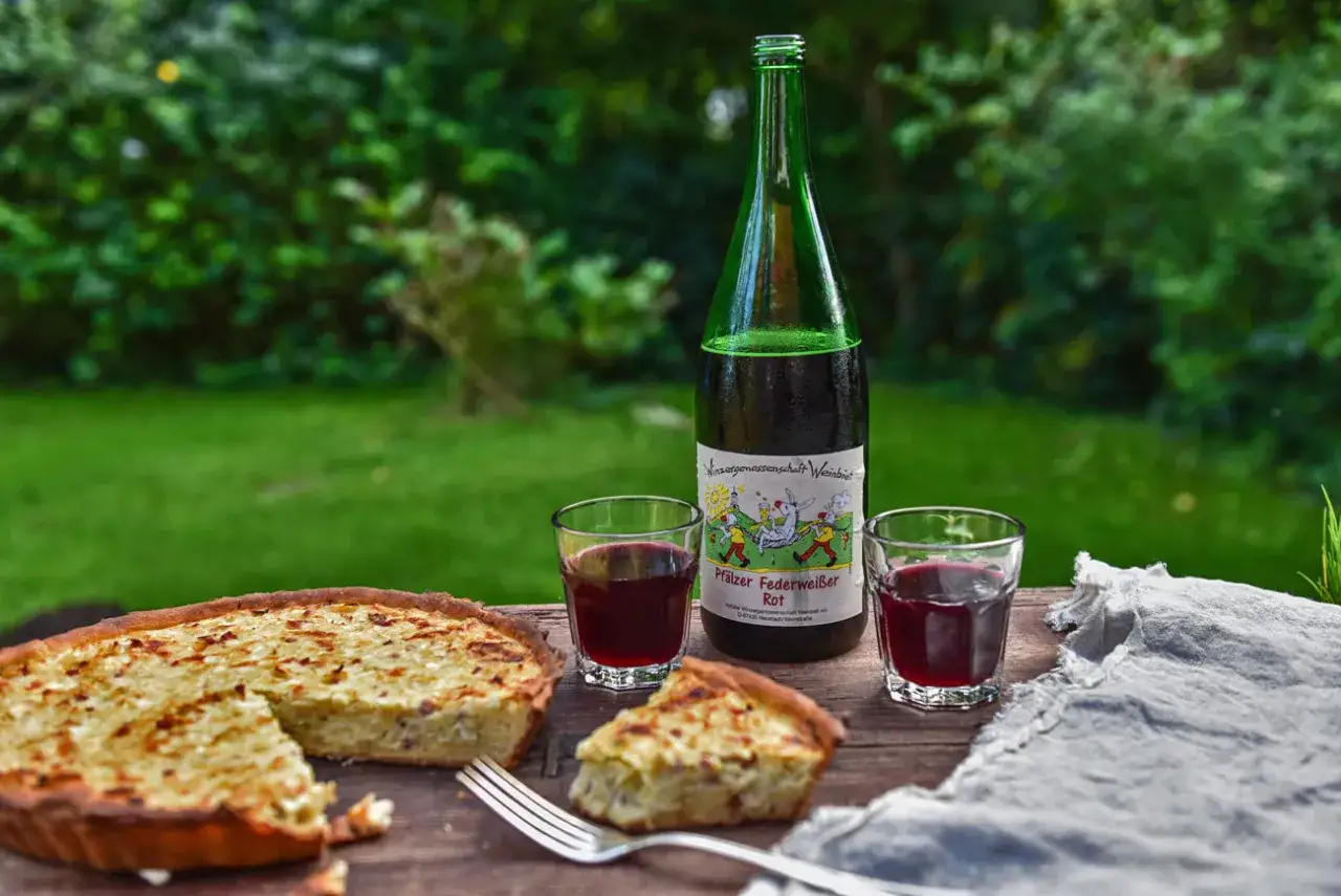 Ein herzhafter Zwiebelkuchen und zwei Gläser Federweißer, der erste Wein im Jahr, stehen auf einem Holztisch im Garten.
