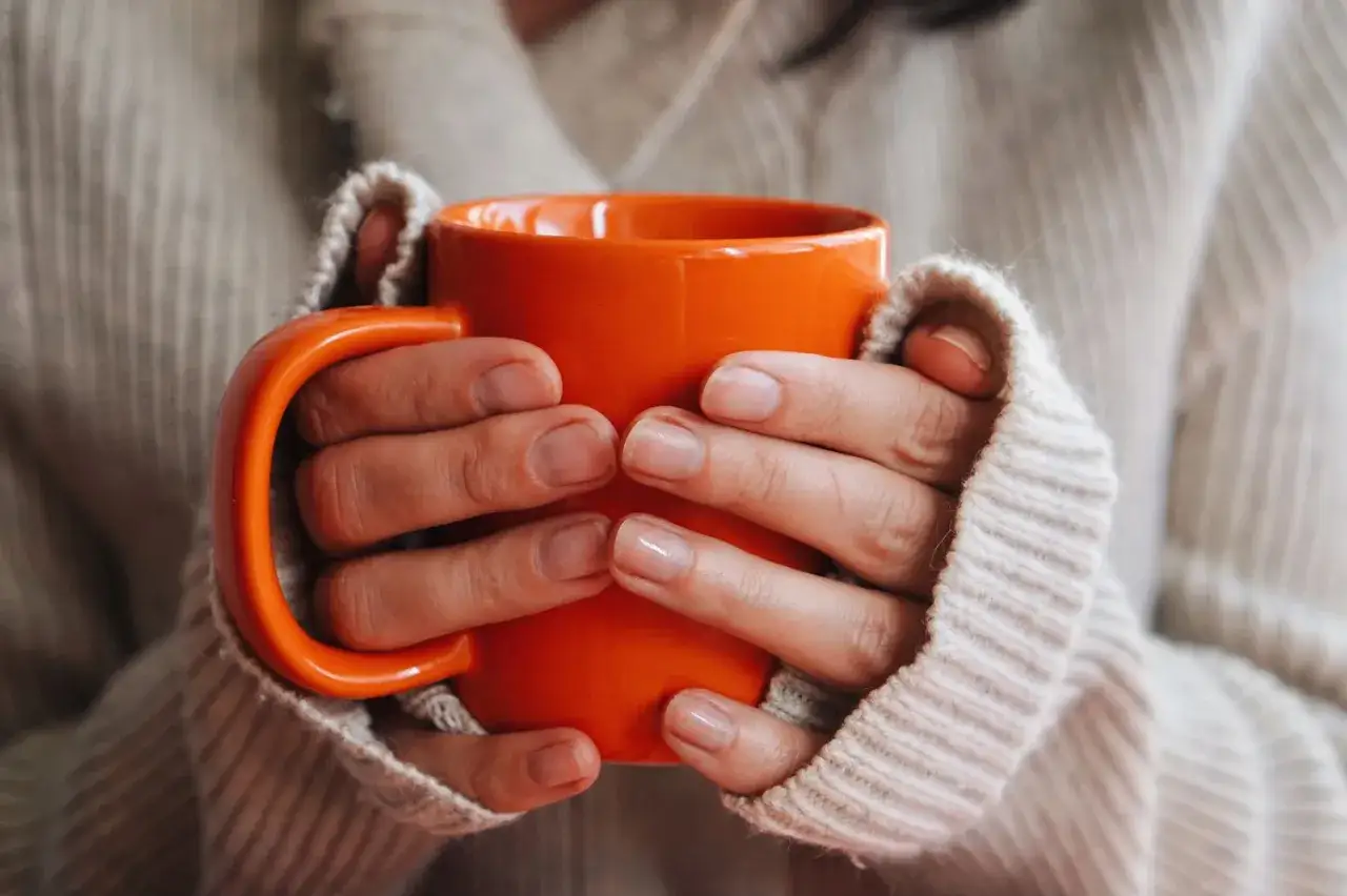 Hände halten eine orangefarbene Tasse mit warmem Getränk, ideal bei Halsschmerzen.